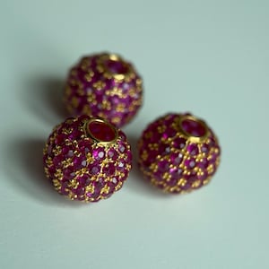 May include: Three round, ornate beads. Each bead is covered in small, vibrant pink gemstones set in a gold-colored metal. The beads are arranged on a white surface, creating a visually appealing display of jewelry.