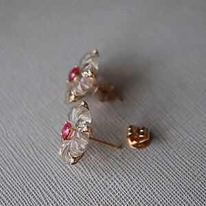 May include: A pair of gold earrings with a clear flower design and a red gemstone in the center.
