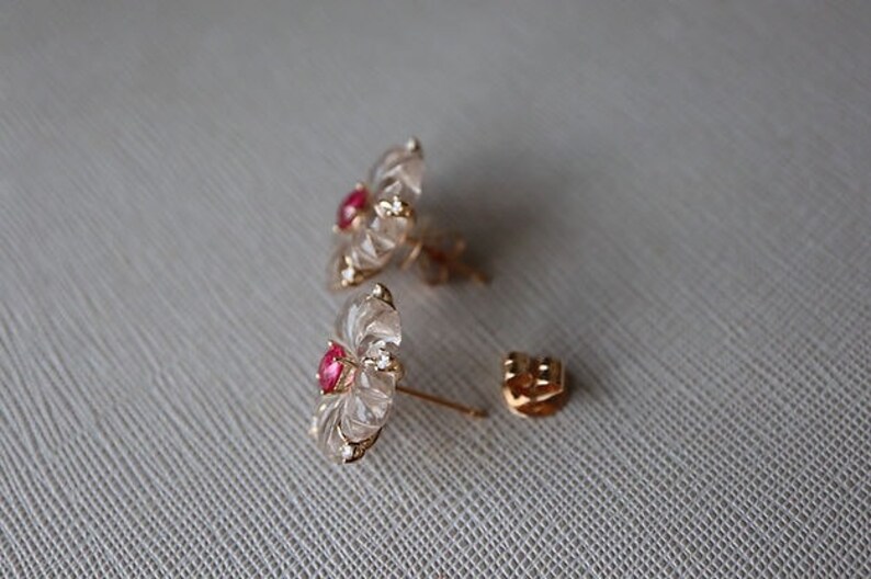 May include: A pair of gold earrings with a clear flower design and a red gemstone in the center.