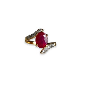 May include: A gold ring featuring a large, oval-cut ruby gemstone. The ring's band has a swirling design, accented with small, round diamonds. The ruby is a deep red color, and the diamonds sparkle against the gold.
