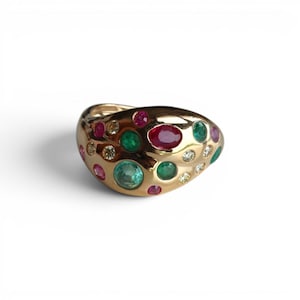 May include: A gold ring featuring a smooth, rounded surface embedded with a variety of colorful gemstones. The ring includes multiple round and oval-shaped rubies, emeralds, and small white diamonds set flush into the polished yellow gold band.