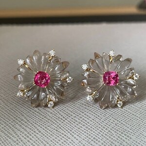 May include: A pair of earrings featuring a clear flower design with a pink gemstone in the center of each flower. The earrings are accented with small, clear gemstones.