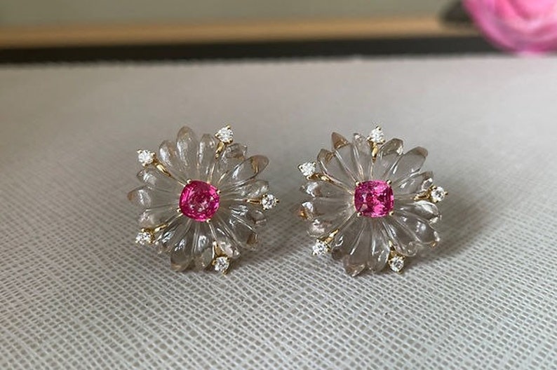 May include: A pair of earrings featuring a clear flower design with a pink gemstone in the center of each flower. The earrings are accented with small, clear gemstones.