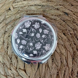 May include: A silver compact mirror with a black and white floral design. The design features white roses and leaves on a black background.