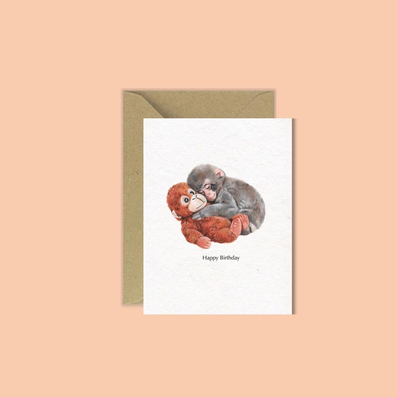 Op de afbeelding: Een wenskaart met een aquarel illustratie van twee baby aapjes die elkaar omhelzen. E&eacute;n aapje is roodbruin, de andere is grijs. De woorden "Happy Birthday" staan eronder gedrukt. De kaart wordt geleverd met een bruine envelop.