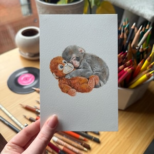 Op de afbeelding: Een aquarel schilderij van twee apen die elkaar omhelzen. E&eacute;n aap is oranje en de andere is grijs. Het kunstwerk staat op een witte rechthoekige kaart, vastgehouden door een persoon. Gekleurde potloden en een plaat staan op de achtergrond.
