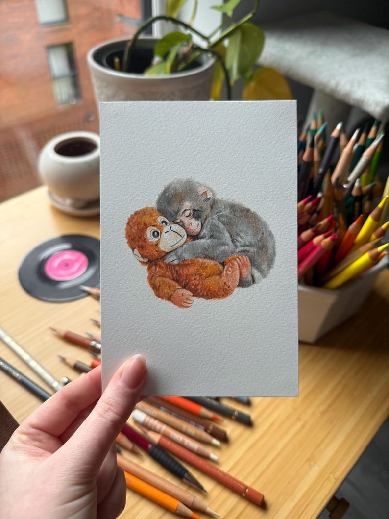 Op de afbeelding: Een aquarel schilderij van twee apen die elkaar omhelzen. E&eacute;n aap is oranje en de andere is grijs. Het kunstwerk staat op een witte rechthoekige kaart, vastgehouden door een persoon. Gekleurde potloden en een plaat staan op de achtergrond.