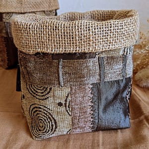 Peut inclure: Un panier de rangement carré en toile de jute et tissu. Le panier a un motif patchwork brun et beige avec une bordure brun foncé. Le panier est posé sur une surface brune.