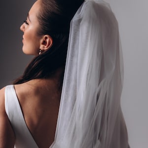 Puede incluir: Una novia con un vestido de novia blanco y un velo largo, sosteniendo un ramo de rosas blancas y follaje verde.