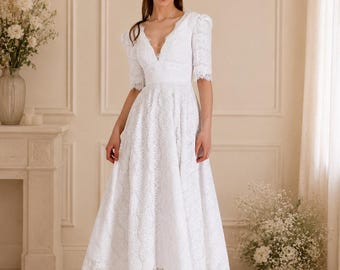 Può includere: Abito midi in pizzo bianco con scollo a V e maniche corte. L'abito presenta un corpetto aderente e una gonna svasata. La modella indossa sandali rosa chiaro. Lo sfondo comprende un camino e composizioni floreali.
