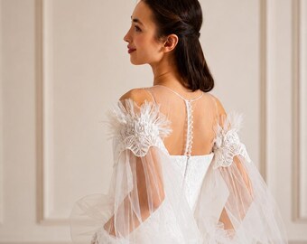 May include: A white floor-length gown featuring sheer tulle bishop sleeves with lace and feather-like trim at the shoulders. The bodice includes a sheer illusion neckline, a vertical row of small buttons along the center back, and a solid skirt.