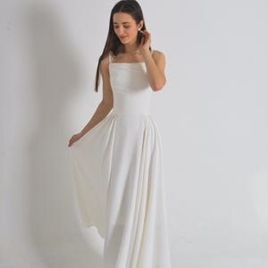 Puede incluir: Un vestido blanco largo con escote cuadrado y tirantes finos. El vestido tiene un corpiño ajustado y una falda fluida. La modelo lleva tacones beige. El vestido es adecuado para bodas y eventos formales.