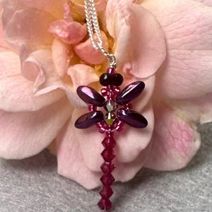 May include: A handmade dragonfly pendant necklace. The dragonfly has iridescent purple wings and a red beaded body. The necklace has a silver chain, set against a soft pink flower.