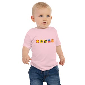 May include: A pink baby t-shirt with a row of five colorful nautical signal flags printed on the front. The flags are red, yellow, black, blue, and orange.
