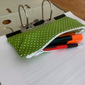 May include: A green polka dot pencil case with a white zipper, holding pens and markers. The case is attached to a binder with silver metal rings. The pens include orange, black, and red colors.