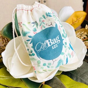 May include: A white cotton drawstring bag with a teal blue circle that says "Gift Bag 123". The bag has a floral pattern with green leaves, red berries, and white cotton bolls.