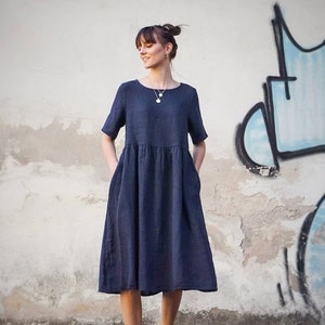 May include: A woman wearing a navy blue linen dress with short sleeves and pockets. She is standing in front of a wall with graffiti and wearing white trainers.