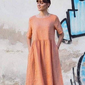 May include: A peach-coloured linen dress with short sleeves and pockets. The dress has a loose, flowing fit and falls to the knees.