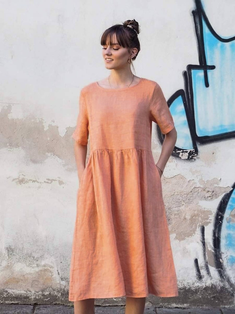 May include: A peach-coloured linen dress with short sleeves and pockets. The dress has a loose, flowing fit and falls to the knees.