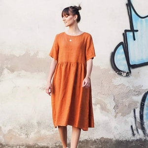 May include: A woman wearing a rust-coloured linen dress with short sleeves and a gathered skirt. She is wearing white trainers and has her hair pulled back in a bun.