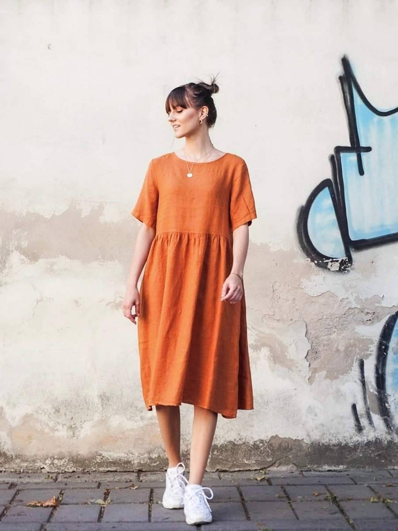 May include: A woman wearing a rust-coloured linen dress with short sleeves and a gathered skirt. She is wearing white trainers and has her hair pulled back in a bun.