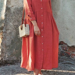 May include: A long-sleeved, red button-down shirt dress with a collared neckline. The dress has a relaxed fit and a slightly gathered skirt. The model is wearing white sneakers and carrying a woven straw bag.