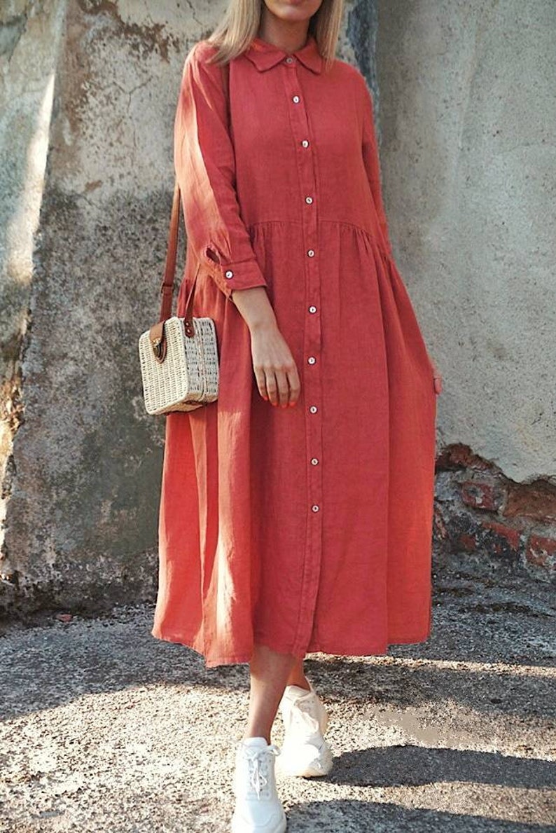 May include: A long-sleeved, red button-down shirt dress with a collared neckline. The dress has a relaxed fit and a slightly gathered skirt. The model is wearing white sneakers and carrying a woven straw bag.