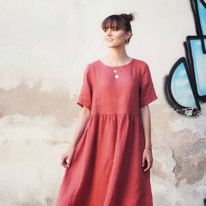 May include: A woman wearing a coral linen dress with short sleeves. The dress has a gathered waist and a midi length.