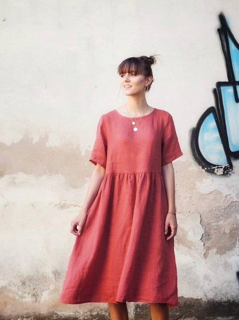 May include: A woman wearing a coral linen dress with short sleeves. The dress has a gathered waist and a midi length.