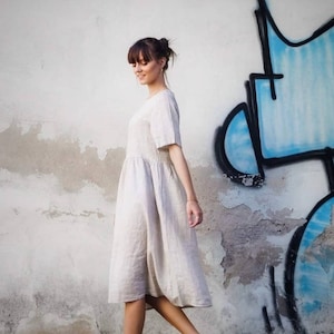 May include: A woman wearing a beige linen dress and white trainers walks in front of a white wall with blue and black graffiti.