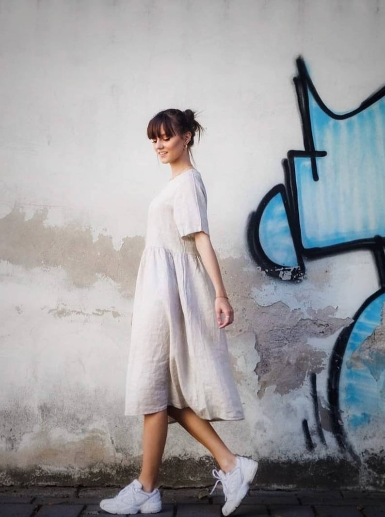 May include: A woman wearing a beige linen dress and white trainers walks in front of a white wall with blue and black graffiti.