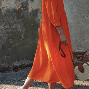 May include: A woman wearing a bright orange long-sleeved dress with a button-up collar and a white straw bag with a brown leather strap.