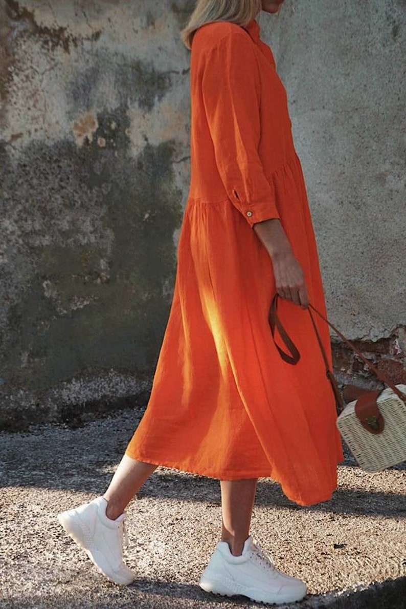 May include: A woman wearing a bright orange long-sleeved dress with a button-up collar and a white straw bag with a brown leather strap.