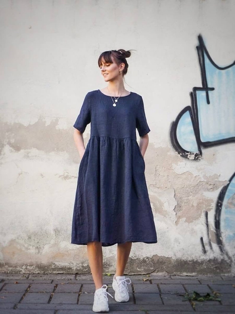 May include: A woman wearing a navy blue linen dress with short sleeves and pockets. She is standing in front of a wall with graffiti and wearing white trainers.