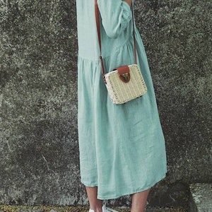 May include: A light blue linen dress with a brown leather strap and a woven straw bag with a brown leather flap and clasp. The person is wearing white sneakers.
