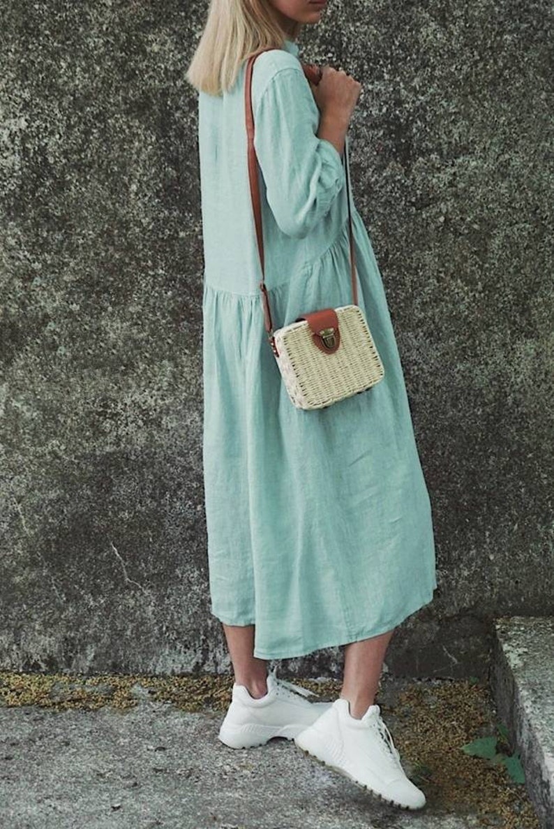 May include: A light blue linen dress with a brown leather strap and a woven straw bag with a brown leather flap and clasp. The person is wearing white sneakers.