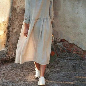 May include: A woman wearing a light beige linen dress with a gathered skirt and 3/4 sleeves. She is walking away from the camera and carrying a brown and green woven bag.