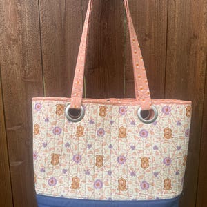 May include: Quilted tote bag with a floral and dog pattern in peach, purple, and cream. The bag has a blue base and handles, with silver grommets. The handles are peach with a blue accent.