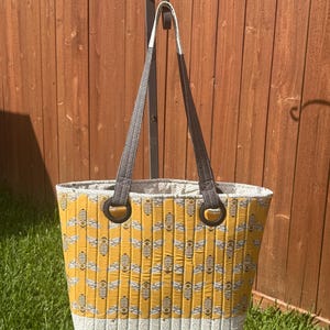 May include: A yellow tote bag with a bee pattern. The bag has a white and gray patterned bottom and a gray and brown handle. The bag is made of quilted fabric and has a rectangular shape.