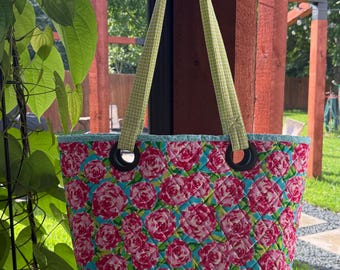 May include: Quilted tote bag with a floral rose pattern in pink and teal. The bag has a green checkered trim and handles, with black grommets for the handles. The bag is hanging outside.
