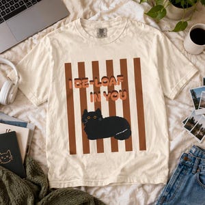 Op de afbeelding: Een crèmekleurig T-shirt met korte mouwen en een afbeelding van een zwarte kat in een liggende houding. Het ontwerp bevat verticale bruine strepen en de tekst "I BE-LOAF IN YOU" in een dik, gelaagd lettertype op de voorkant van het shirt.