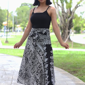 May include: A woman wearing a black tank top and a long, black and white patterned skirt. She is standing on a paved path in a park.