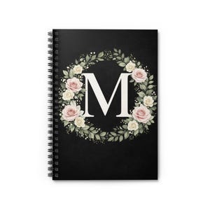 Könnte beinhalten: Ein spiralgebundenes Notizbuch mit schwarzem Einband. Der Einband zeigt einen weißen Buchstaben "M", umgeben von einem Kranz aus rosa und cremefarbenen Rosen und grünen Blättern. Zum Notieren.