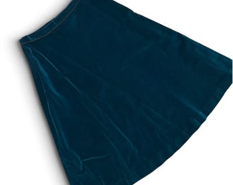 Könnte beinhalten: Ein tief petrolfarbener Samtrock mit hohem Bund und ausgestellter Silhouette. Der Stoff hat eine weiche, lichtreflektierende Textur mit glatter Oberfläche, die von der Taille bis zu einem geraden Saum in einem einfarbigen Blaugrün verläuft.
