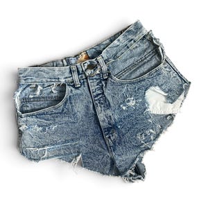 Peut inclure: Short en jean bleu délavé à l'acide avec une taille haute, une fermeture à bouton et à glissière, des passants de ceinture et plusieurs zones usées aux bords effilochés. Le short présente une déchirure marquée sur le côté gauche.
