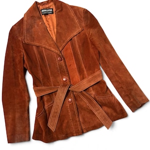 May include: A rust-colored suede leather jacket featuring a wide collar, long sleeves, and a matching tie-waist belt. The front closure includes three buttons, and the interior label reads Genuine Leather, made in Uruguay.