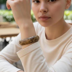 May include: A wide, cylindrical bracelet with a snakeskin pattern. The bracelet is primarily brown with black and tan markings. The image shows the bracelet on a person's wrist, with a neutral background.
