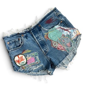 Peut inclure: Short en jean bleu avec un ourlet effiloché, orné de divers empiècements en tissu et de broderies. Un écusson bien visible en bas à gauche porte l'inscription "I Will If You Will" en rouge et vert. Le vêtement présente des détails usés et des coutures.