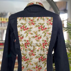 Può includere: Una giacca blu navy con un pannello ricamato floreale sulla schiena e sul colletto. Il pannello presenta fiori rosa e foglie verdi su uno sfondo chiaro. La giacca è esposta su una gruccia.