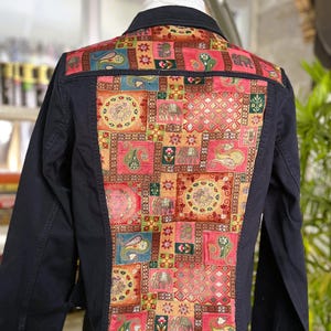 May include: A navy blue denim jacket with a colorful patchwork design on the back. The patchwork features squares with paisley, floral, and elephant motifs in shades of pink, orange, green, and blue. The jacket has long sleeves and a collar.
